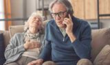 En ældre mand med briller ser bekymret ud, mens han taler i telefon, muligvis med lægevagten, mens han sidder ved siden af en ældre kvinde, der læner sig tilbage med hånden på brystet og ser ud til at have det dårligt.