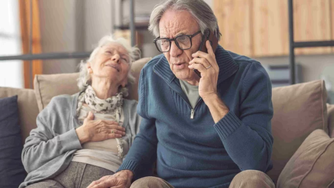 En ældre mand med briller ser bekymret ud, mens han taler i telefon, muligvis med lægevagten, mens han sidder ved siden af en ældre kvinde, der læner sig tilbage med hånden på brystet og ser ud til at have det dårligt.
