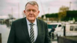 En midaldrende mand i mørkt jakkesæt og stribet slips står udendørs ved en lystbådehavn med både og vand i baggrunden på en overskyet dag, der minder om Lars Løkke under regeringsforhandlinger mellem V og K.