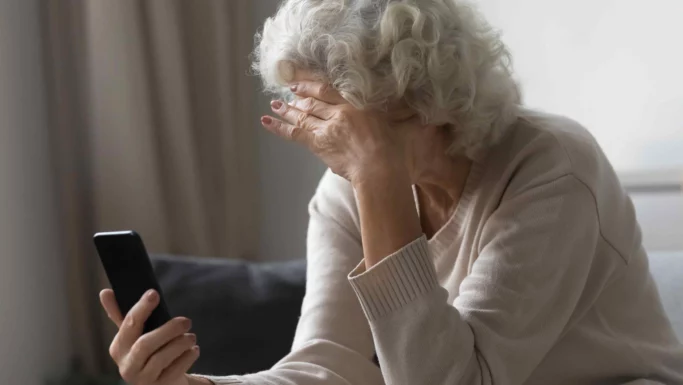 En ældre kvinde med gråt hår sidder i en sofa, holder en smartphone i den ene hånd og dækker sit ansigt med den anden, og hun ser fortvivlet ud - måske søger hun hjælp efter kriminalitet eller hjælp efter ulykker.