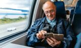 En ældre mand med briller sidder ved et togvindue og bruger sin smartphone med brun etui. Iført en blå jakke er han fokuseret på sin enhed, mens landskabet glider forbi - et øjebliksbillede af seniorer på rejser.