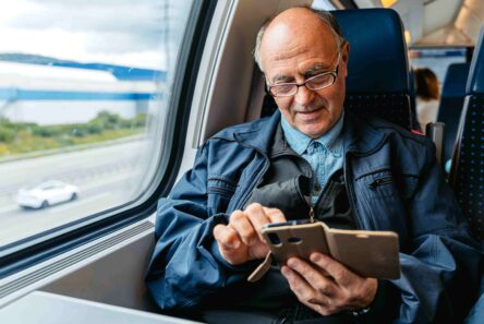 En ældre mand med briller sidder ved et togvindue og bruger sin smartphone med brun etui. Iført en blå jakke er han fokuseret på sin enhed, mens landskabet glider forbi - et øjebliksbillede af seniorer på rejser.