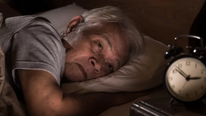 En ældre mand ligger i sengen, ser bekymret ud og kan ikke sove, sover dårligt med hovedet på en pude og et vækkeur, der viser 2:59 på natbordet ved siden af ham.