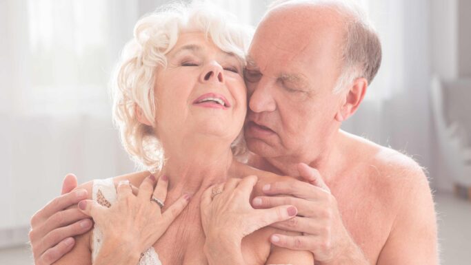 Et ældre par omfavner hinanden intimt, mens manden forsigtigt holder om kvinden bagfra. Begge ser ud til at nyde et ømt, kærligt øjeblik sammen, hvilket fremhæver varmen og nærheden i deres sexliv i et blødt oplyst rum.