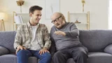 En ung mand og en ældre mand sidder sammen i en sofa, smiler og taler om arveret. Den ældre mand gestikulerer, mens han fortæller en historie. Rummet er hyggeligt med hylder og dekorative planter i baggrunden.