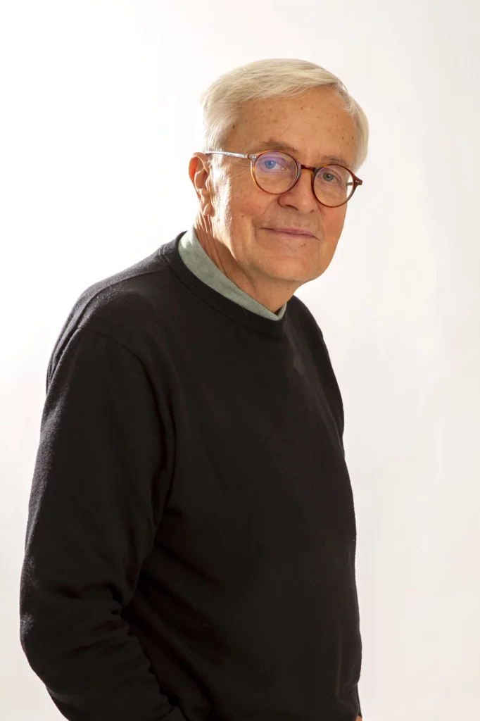 En ældre mand med kort hvidt hår og briller, iført en sort sweater over en grøn skjorte, står mod en almindelig lys baggrund og kigger på kameraet med et lille smil - måske klar til at finde kærligheden på Hotel Romantik for singler.