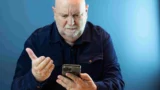 En ældre mand med skæg og mørkeblå skjorte ser forvirret ud, mens han holder en smartphone, frustreret over det, der måske er et skjult abonnement, på en almindelig blå baggrund.