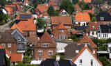 Et tæt kvarter med traditionelle europæiske huse med stejle, røde og mørke tage, murstensvægge og grønne træer spredt mellem husene på en solrig dag - et område, hvor Alka-kunder sætter pris på deres forsikring for at få ro i sindet.