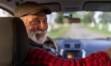 En ældre mand med gråt skæg, kasket og rødternet skjorte smiler, mens han sidder på førersædet i en bil og afspejler den tilfredshed, som mange bilejere føler med deres betroede bilmærker, når han ser sig tilbage over skulderen. Vejen forude er synlig gennem forruden.