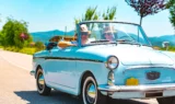 Et ældre par iført solbriller og hatte kører i en lyseblå vintage-cabriolet ned ad en solrig landevej og nyder bilferien med grønne træer og bjerge i baggrunden.
