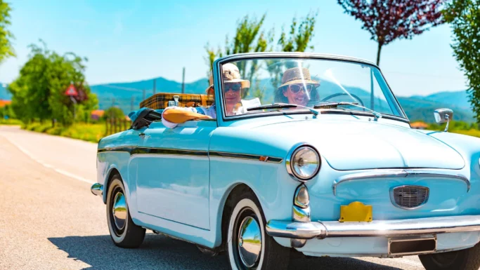 Et ældre par iført solbriller og hatte kører i en lyseblå vintage-cabriolet ned ad en solrig landevej og nyder bilferien med grønne træer og bjerge i baggrunden.