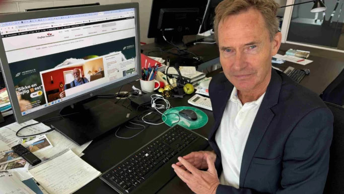 En mand i jakkesæt sidder ved et skrivebord med rodede papirer og kontorartikler. Med front mod kameraet viser hans store skærm en hjemmeside om ChatGPT-abonnement, hvilket antyder, at han måske er ved at undersøge, hvordan man undgår at blive snydt eller bedraget online.