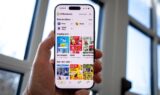 En hånd holder en smartphone med tilbud på skærmen, hvor forskellige butiklogos og produktbilleder vises. I baggrunden strømmer dagslys ind gennem store vinduer.