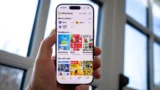 En hånd holder en smartphone med tilbud på skærmen, hvor forskellige butiklogos og produktbilleder vises. I baggrunden strømmer dagslys ind gennem store vinduer.