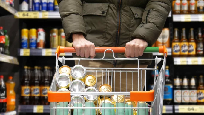 En person i en grøn jakke skubber en indkøbsvogn fyldt med pantfri dåser i en butiksgang med flasker og dåser, hvilket understreger grænsehandelens tiltrækningskraft.