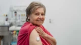 En ældre kvinde smiler, mens hun viser sin overarm med en bandage efter at have fået en vaccine mod helvedesild, siddende i et medicinsk miljø med en hvid baggrund.