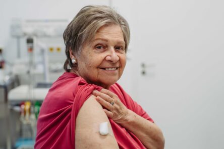 En ældre kvinde smiler, mens hun viser sin overarm med en bandage efter at have fået en vaccine mod helvedesild, siddende i et medicinsk miljø med en hvid baggrund.