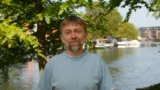 En mand med lysebrunt hår og skæg, iført en lyseblå t-shirt, står udendørs ved en flod med træer, både og bygninger i baggrunden på en solrig dag - perfekt til en guide til, hvordan man kommer i gang med aktiesparekonto.