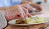Nærbillede af en ældre persons hånd, der holder en gaffel og kniv, mens hun nyder hjemmelavet mad med kartoffelmos på et plejehjem, mens andre mennesker spiser i baggrunden.