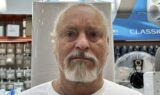 Jan Gindrup står indendørs og kigger ind i kameraet. Han er iført en hvid skjorte og en guldkæde. Bag ham antyder hylder og en stor ventilator, at han befinder sig i en butik, hvor han muligvis diskuterer pensions- eller tjenestemandspensionsspørgsmål.