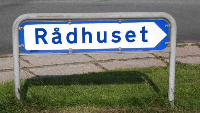 Et gadeskilt med ordet Rådhuset i blå bogstaver på en hvid baggrund med blå kant peger til højre - måske leder det dig mod samtaler om politikerne løn. Skiltet er monteret på metalskinner og står på græs ved siden af et fortov.