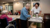 En plejer serverer drikkevarer for en ældre kvinde ved et bord på et kommunalt plejehjem, mens andre ældre sidder og ser på. En kvinde i kørestol står i forgrunden og understreger den rolige, fælles omsorg for sårbare borgere.