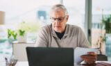 En ældre mand med gråt hår og briller sidder ved et skrivebord og kigger på en bærbar computer, mens han holder papirer om opsparing. Planter og store vinduer lyser op i den moderne baggrund, mens han overvejer, om han skal investere i aktier.