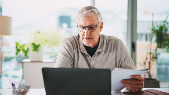 En ældre mand med gråt hår og briller sidder ved et skrivebord og kigger på en bærbar computer, mens han holder papirer om opsparing. Planter og store vinduer lyser op i den moderne baggrund, mens han overvejer, om han skal investere i aktier.