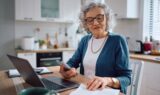 En ældre kvinde med gråt hår og briller sidder ved et køkkenbord med en bærbar computer, en smartphone og papirer og ser ud til at gennemgå sin pensionsopgørelse eller styre økonomien hjemmefra. Hun er iført en hvid skjorte og en blå cardigan.