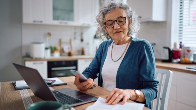 En ældre kvinde med gråt hår og briller sidder ved et køkkenbord med en bærbar computer, en smartphone og papirer og ser ud til at gennemgå sin pensionsopgørelse eller styre økonomien hjemmefra. Hun er iført en hvid skjorte og en blå cardigan.