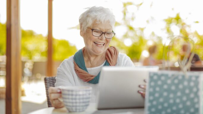 En ældre kvinde med kort, hvidt hår og briller sidder ved et udendørs bord og smiler til en tablet. Hun holder et krus i den ene hånd - måske tjekker hun tips til pensionister for at spare penge - mens hun nyder en solrig dag. En blå og hvid prikket taske står på bordet.