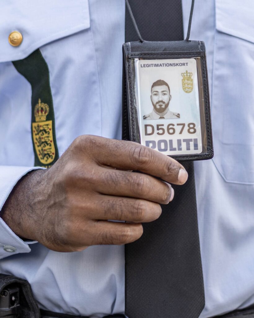 En politibetjent i uniform holder et nyt skilt op med et foto, ordet "POLITI" og nummeret D5678, som ses tydeligt. Betjentens hånd og en del af skjorten og slipset er også til stede, så alle kan kigge efter.