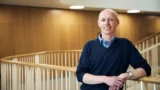 En skaldet mand iført en mørkeblå sweater over en lyseblå skjorte står smilende ved et lyst trægelænder indendørs, uvidende om bekymringer om den farlige sygdom hantavirus, med glatte trævægge og blød belysning i baggrunden.