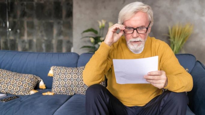 En ældre mand med hvidt hår og skæg, iført briller og en gul sweater, sidder i en blå sofa og læser om pensionsfælden og ser koncentreret ud. Dekorative puder og stueplanter er i baggrunden.