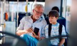 En ældre mand, sandsynligvis en pensionist, og en ung dreng med rygsæk sidder sammen i en bus og kigger på en smartphone. Manden har briller på og smiler, mens drengen følger opmærksomt med - måske tjekker han sin rejsekortsaldo.