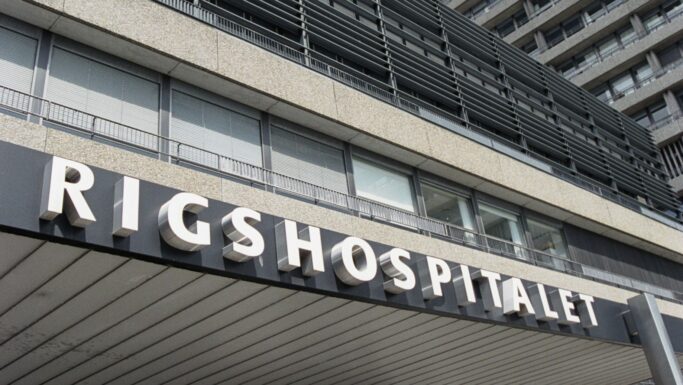 Billedet viser den udvendige indgang til Rigshospitalet, der er kendt for hjertebehandling, med store bogstaver over døråbningen. Bygningen har moderne arkitektur med vandrette vinduer og betonpaneler.