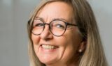 En smilende ældre kvinde med langt, lysebrunt hår, iført runde briller og perleøreringe, vist i nærbillede mod en neutral baggrund - ideel til råd til pårørende om at møde mennesker med demens.