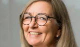 En smilende ældre kvinde med langt, lysebrunt hår, iført runde briller og perleøreringe, vist i nærbillede mod en neutral baggrund - ideel til råd til pårørende om at møde mennesker med demens.