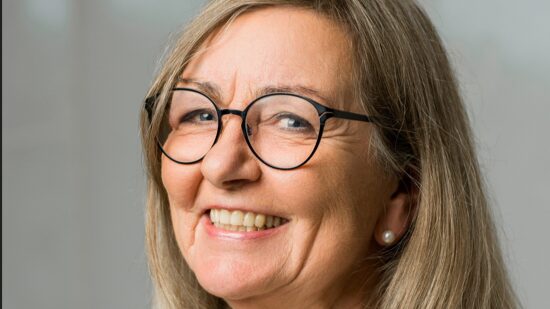 En smilende ældre kvinde med langt, lysebrunt hår, iført runde briller og perleøreringe, vist i nærbillede mod en neutral baggrund - ideel til råd til pårørende om at møde mennesker med demens.