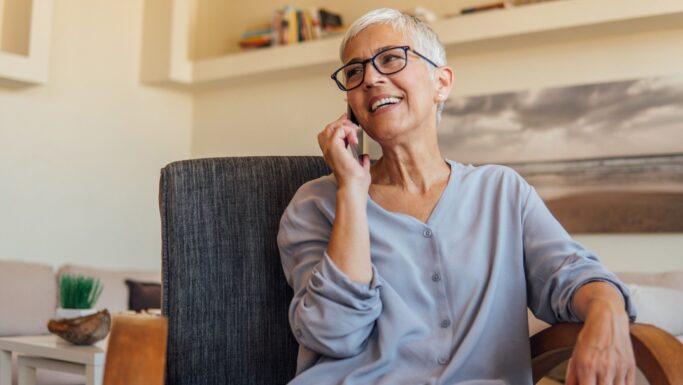 En ældre kvinde med kort gråt hår og briller sidder i en stue, smiler og taler i en smartphone. Hun har en lyseblå bluse på og virker afslappet, uvidende om mulige svindel fra falske politifolk. Hylder og et landskabsfoto er i baggrunden.