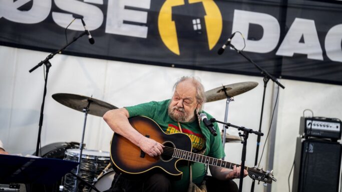 Stig Møller, en ældre mand med skæg, spiller akustisk guitar og synger i en mikrofon på scenen, med trommer og forstærkere bag sig og et stort eventbanner i baggrunden, og fanger en anden generation.