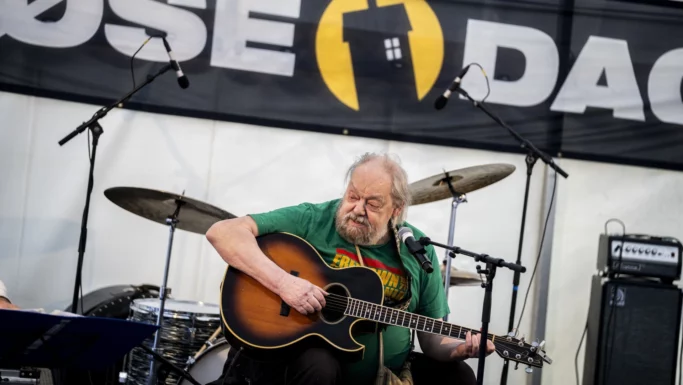 Stig Møller, en ældre mand med skæg, spiller akustisk guitar og synger i en mikrofon på scenen, med trommer og forstærkere bag sig og et stort eventbanner i baggrunden, og fanger en anden generation.