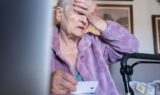 En ældre kvinde i en lilla kappe sidder inden døre og holder sin pande i nød med den ene hånd og et kort - muligvis knyttet til ældre svindel eller manipulation - i den anden. En rollator står ved siden af hende, og indrammede billeder hænger på væggen bag hende.