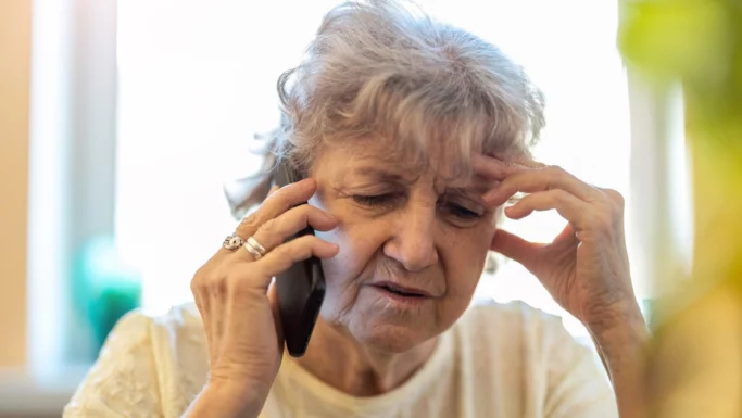 En ældre kvinde med gråt hår holder en telefon op til øret og ser bekymret eller stresset ud, mens hendes anden hånd rører ved hendes pande. Indendørs i en lys top taler hun måske med en svindler, der udgiver sig for at være falsk betjent om hendes smykker.