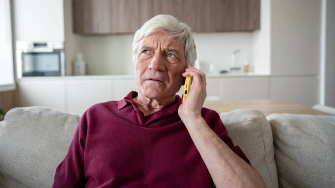 En ældre mand med gråt hår sidder i en sofa i et moderne køkken, iført en rødbrun skjorte og med en gul telefon ved øret, og han ser bekymret ud, mens han lytter intenst til hackerangreb og falske politifolk.