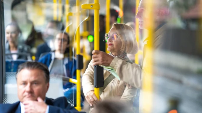 En gruppe mennesker kører i bus; en kvinde med kort blondt hår og briller står og holder en gul pæl, mens andre sidder eller står omkring hende, sløret af refleksioner - måske busser erstatter tog for deres rejse i pinsen.
