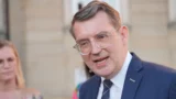 Troels Lund Poulsen, en mand i blåt jakkesæt, hvid skjorte og briller, taler udendørs. Han har kort, brunt hår og kigger lidt til siden. En sløret person står i baggrunden med en bygning bag sig, danne ny regering.