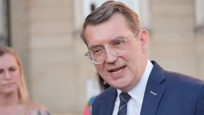 Troels Lund Poulsen, en mand i blåt jakkesæt, hvid skjorte og briller, taler udendørs. Han har kort, brunt hår og kigger lidt til siden. En sløret person står i baggrunden med en bygning bag sig, danne ny regering.