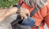 En person bruger et tyveri-trick til i hemmelighed at tage en stjålet punge fra rygsækken på en ældre kvinde med langt gråt hår og en orange top, hvilket tyder på et lommetyveri udendørs.