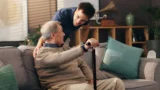 En ung mand smiler og læner sig frem for at tale med en ældre mand, der sidder i en sofa og holder en stok. Det varme, solbeskinnede rum udstråler støtte, mens de diskuterer de seneste nyheder om en person, der er blevet svindlet for 250.000 kroner i den hyggelige stue.